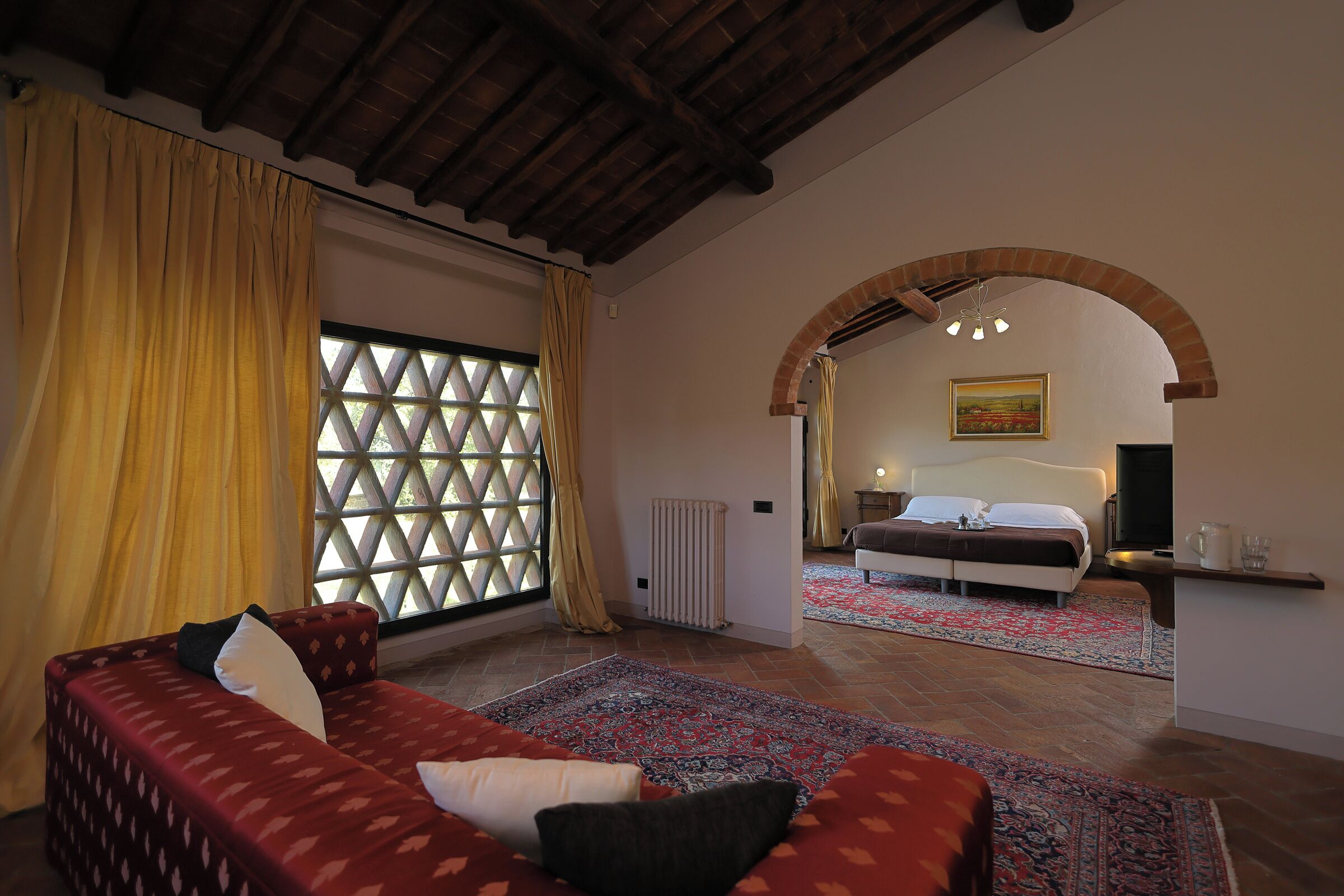 A cozy room with a patterned sofa, a large window with curtains, a patterned rug, and a visible bed in the background.
