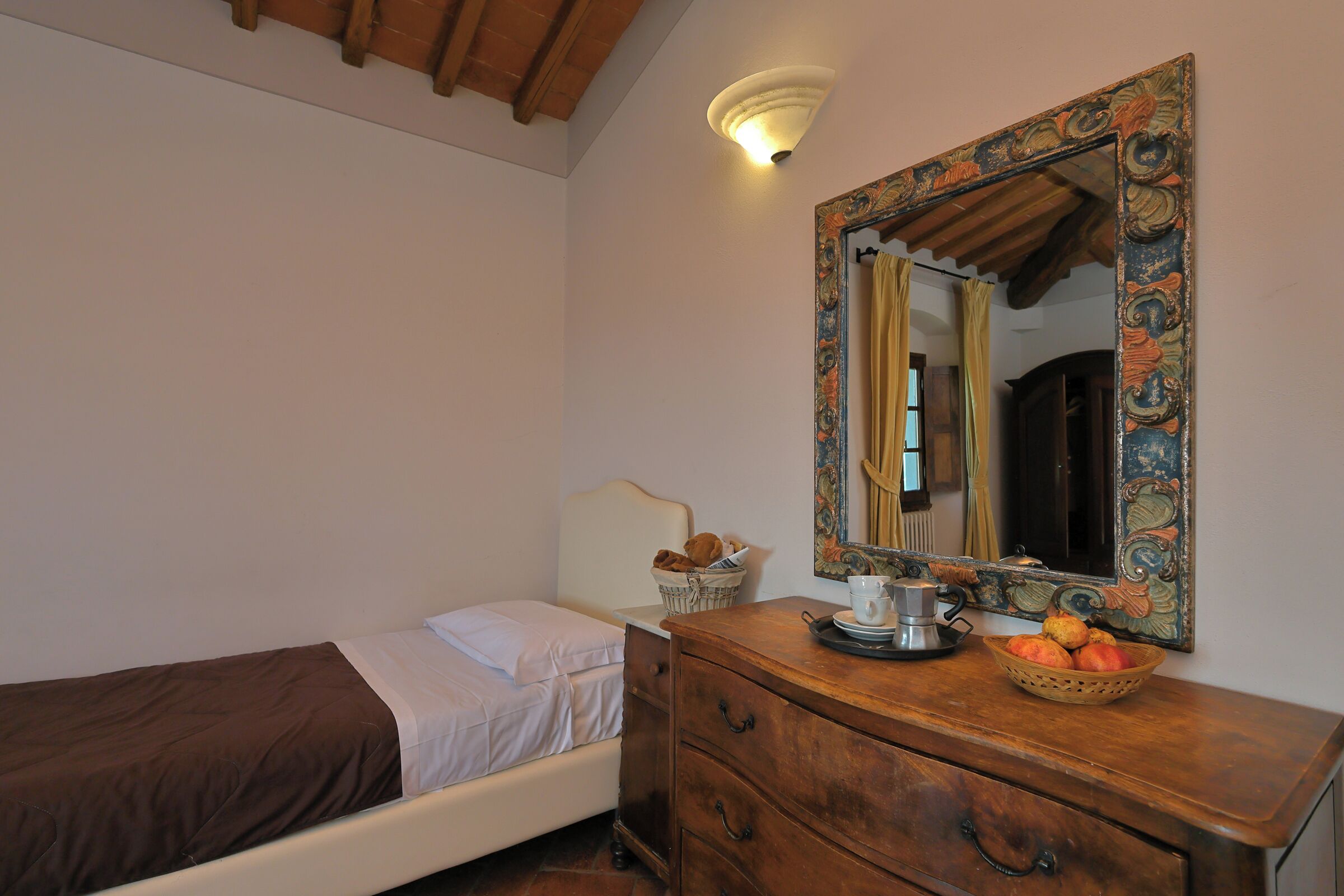 Une chambre à coucher avec un lit simple, une commode en bois, un miroir et un luminaire fixé au mur.