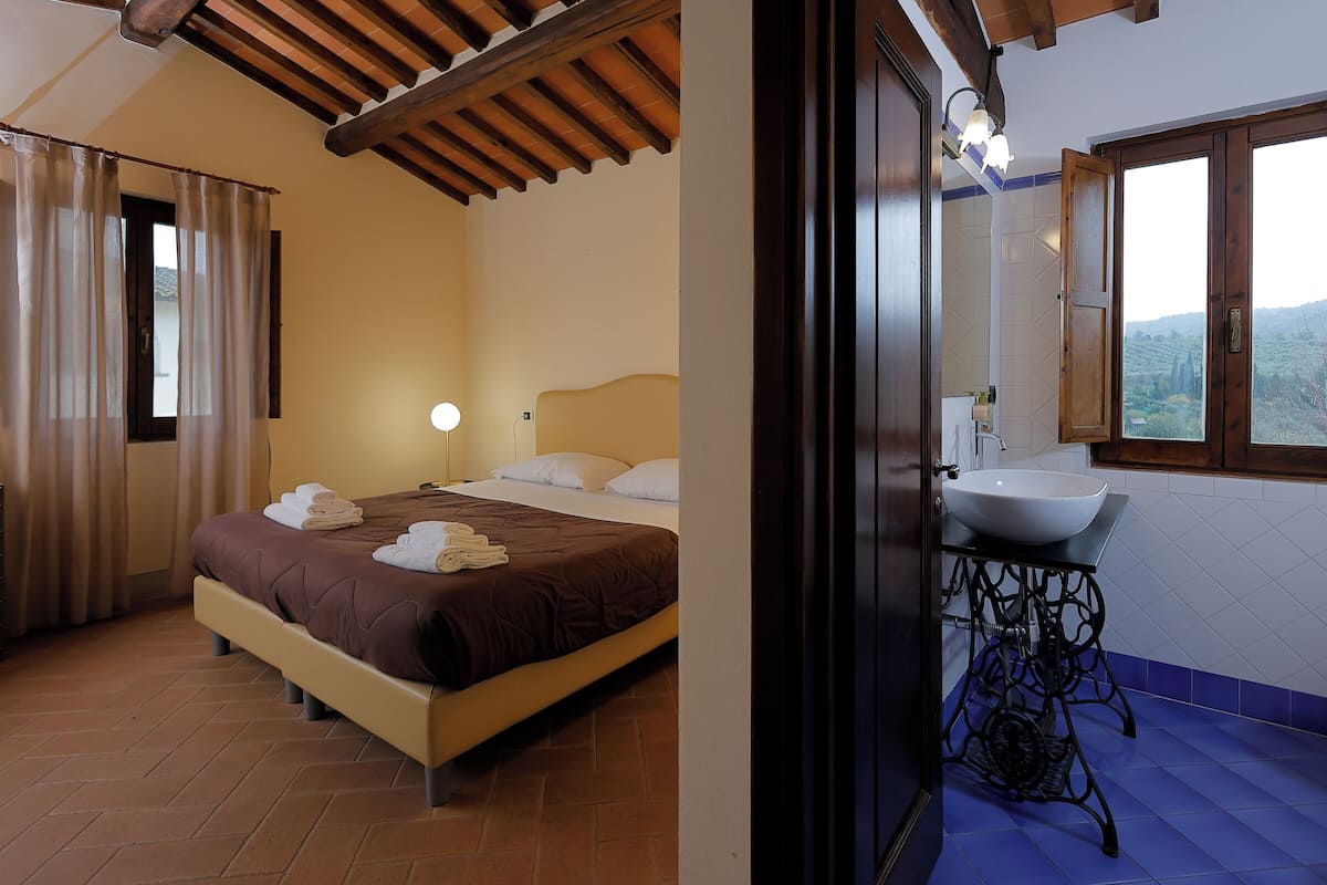 Ein Schlafzimmer mit Bett, Vorhängen und Nachttisch. Es gibt ein Badezimmer mit Waschbecken, Spiegel und Fenster.