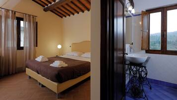 Een slaapkamer met een bed, gordijnen en een nachtkastje. Er is een badkamer met een wastafel, spiegel en raam.
