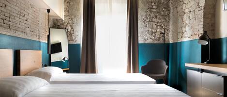 A hotel room with a rustic brick wall, a large bed, a desk with a chair, and a window with curtains.