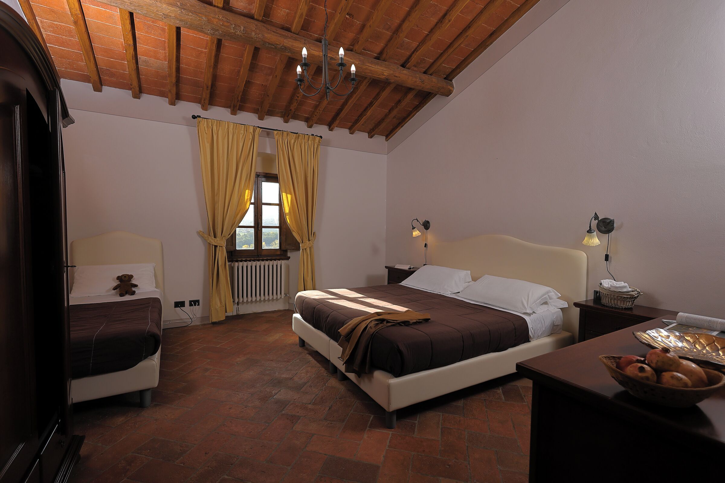 A bedroom with a wooden ceiling, a bed with a headboard, two bedside lamps, a window with curtains, and a small table with a bowl of fruit.