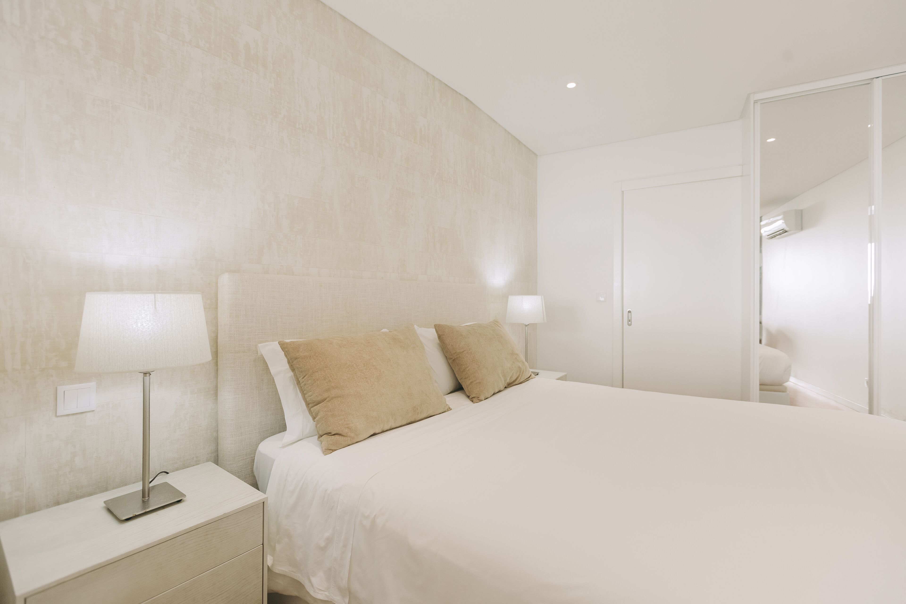 Quarto de hotel moderno com uma cama grande, duas luminárias de cabeceira, uma mesinha de cabeceira e uma porta que dá para outro cômodo.