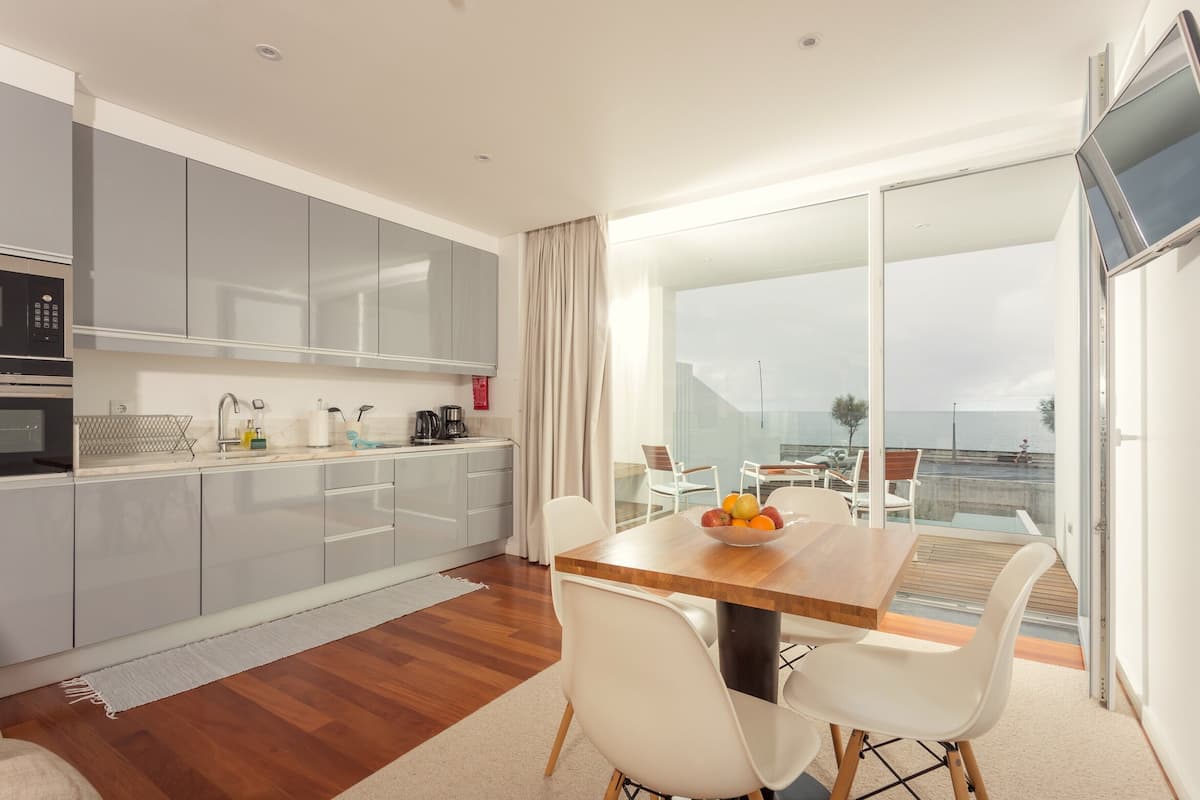Una cocina moderna con armarios grises, una mesa de comedor de madera con sillas blancas y vistas al mar a través de amplios ventanales.