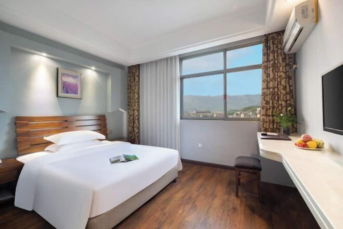 A hotel room with a large bed, a desk with a fruit bowl, a TV, and a window with a view of mountains.