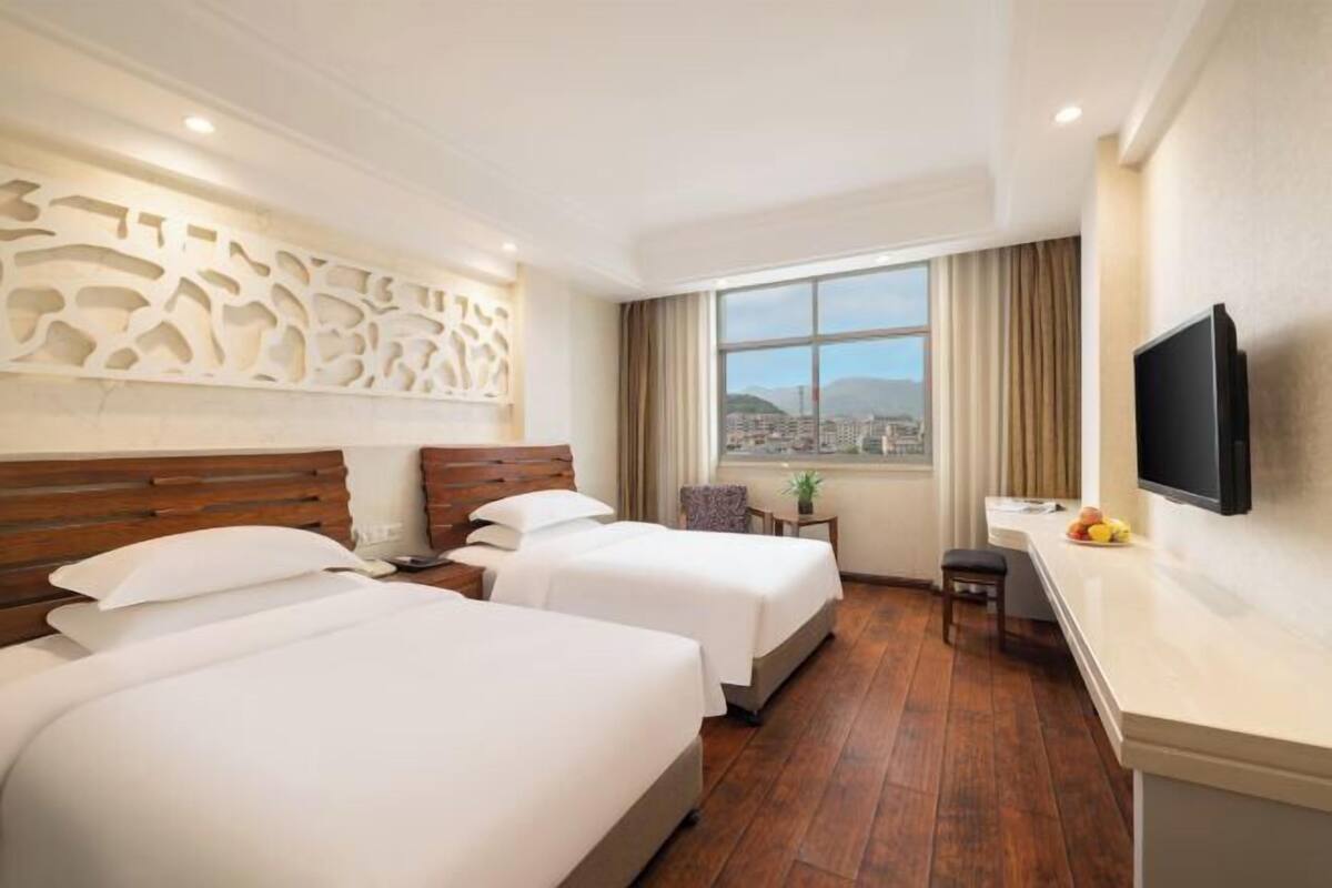 A hotel room with two beds, a TV, a desk, and a window with a city view.