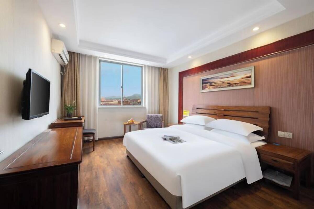 A hotel room with a large bed, a desk with a flat-screen TV, a window with a city view, and a painting on the wall.