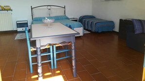 Um quarto pequeno e arrumado com uma cama, uma mesa e uma cadeira. Há uma estante com livros e uma mesinha com uma tigela.