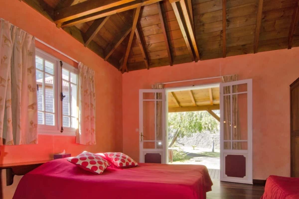 Un dormitorio con techo de madera, cama con ropa de cama roja y vistas a un jardín a través de puertas abiertas.