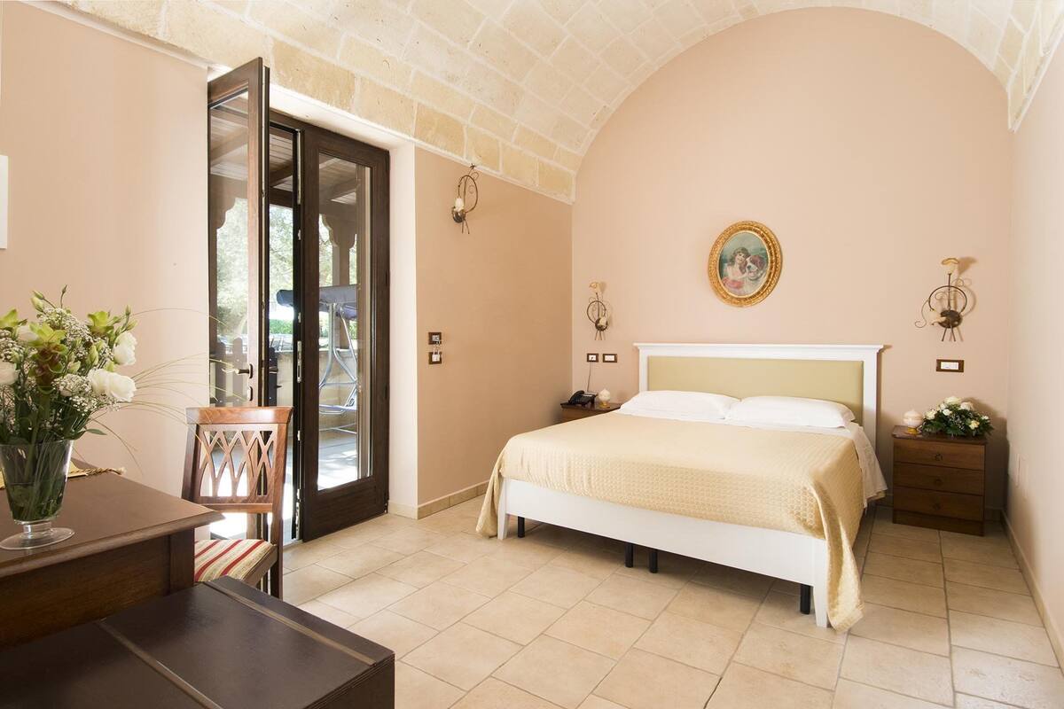 Une chambre à coucher comprenant un lit, des tables de chevet, un bureau et une chaise. Une porte donne accès à un espace extérieur.