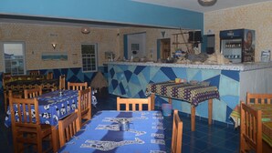 Un restaurante con piso de baldosas azules y beige, mesas y sillas de madera, una zona de bar con refrigerador y una pared decorada con un diseño.