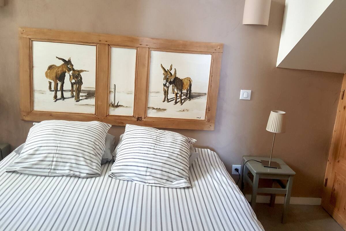 A bedroom with a bed, two bedside tables, a lamp, and a wall-mounted painting of donkeys.