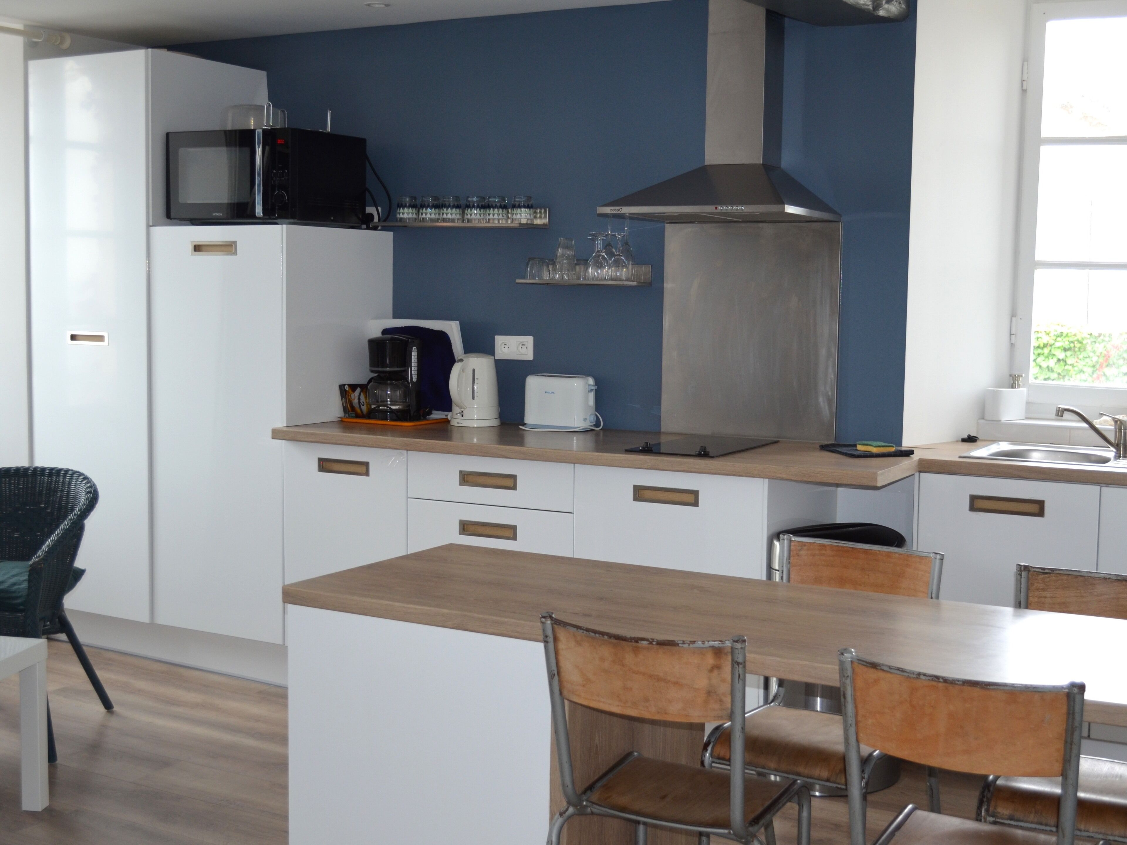 A modern kitchen with a microwave, coffee maker, toaster, and stainless steel range hood. There is a dining table with chairs and a window providing natural light.