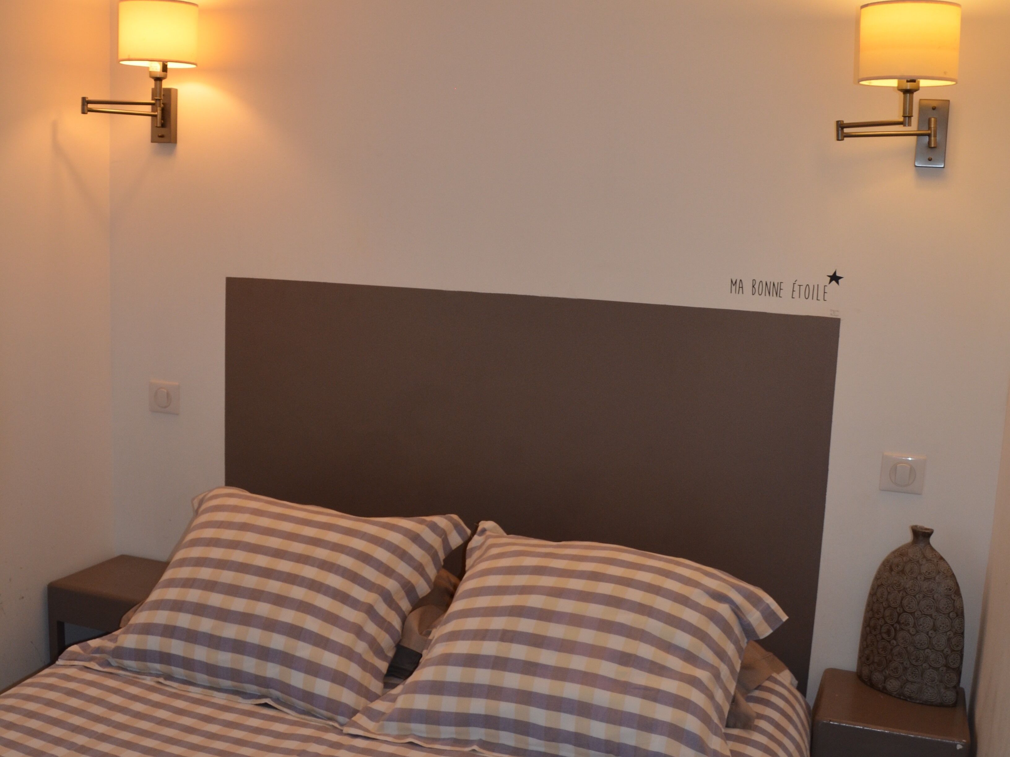 Un lit avec une literie à carreaux, une tête de lit sombre et deux lampes fixées au mur.
