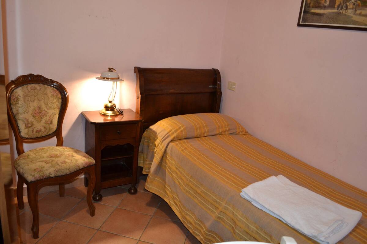A small, neatly arranged bedroom with a single bed, a chair, a nightstand with a lamp, and a wall-mounted painting.