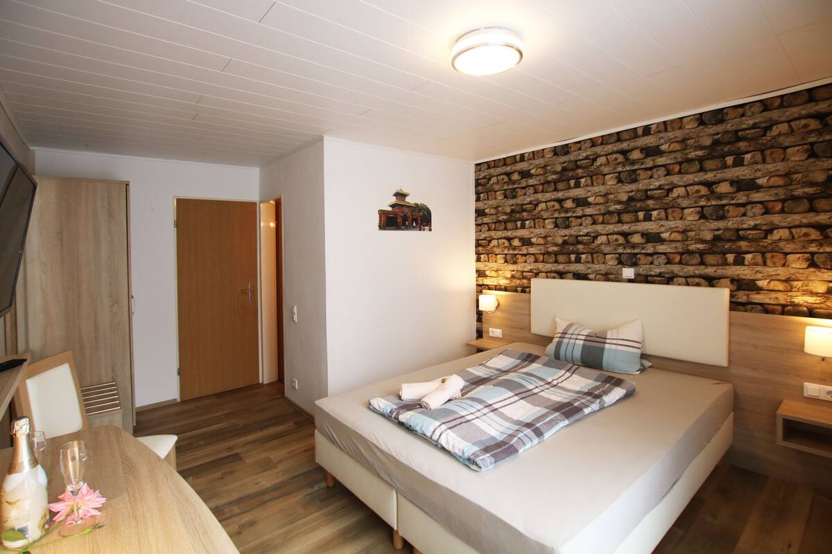 A bedroom with a wooden headboard, a stone wall, a wooden floor, a bed with a plaid blanket, a bedside table with a lamp, and a wooden wardrobe.
