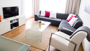 Een moderne woonkamer met een flatscreen televisie, een grijze bank met rode en witte kussens, een glazen salontafel en een witte fauteuil.