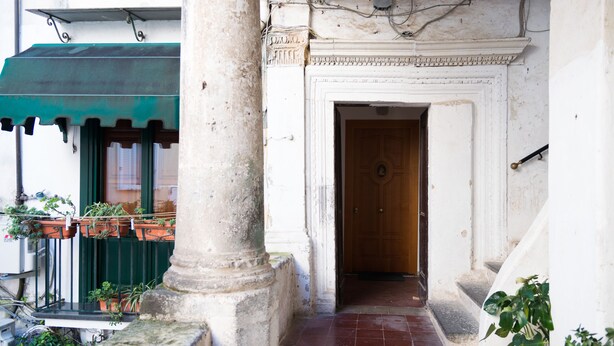 Una entrada estrecha con puerta de madera, suelo de baldosas y toldo verde sobre una ventana.