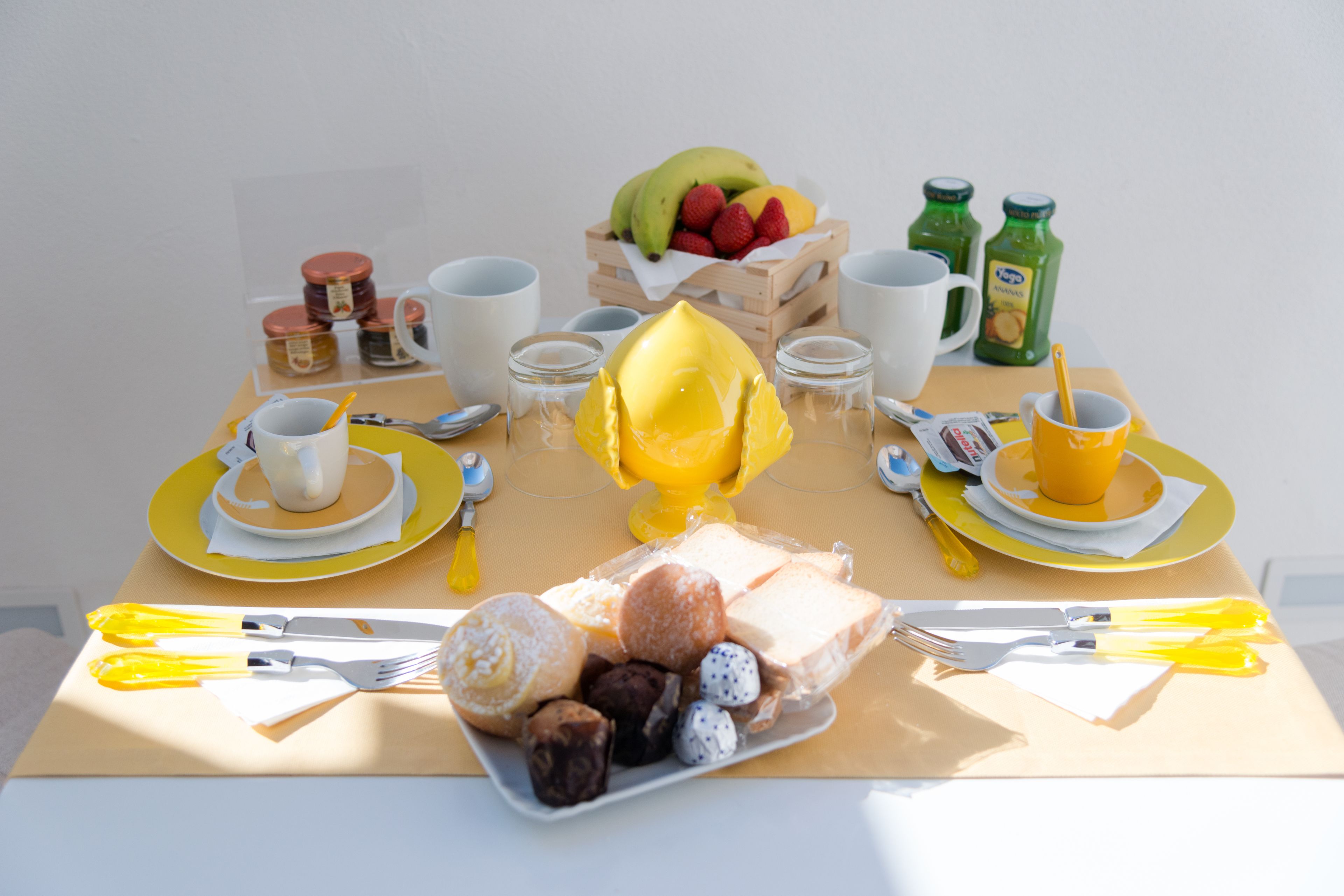 A breakfast table with yellow plates, white cups, and various food items including fruits, pastries, and a glass of orange juice.