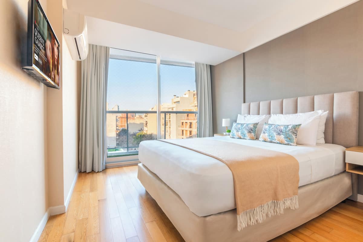 A modern hotel room with a large bed, a flat-screen TV mounted on the wall, a window with curtains, and a view of the cityscape.