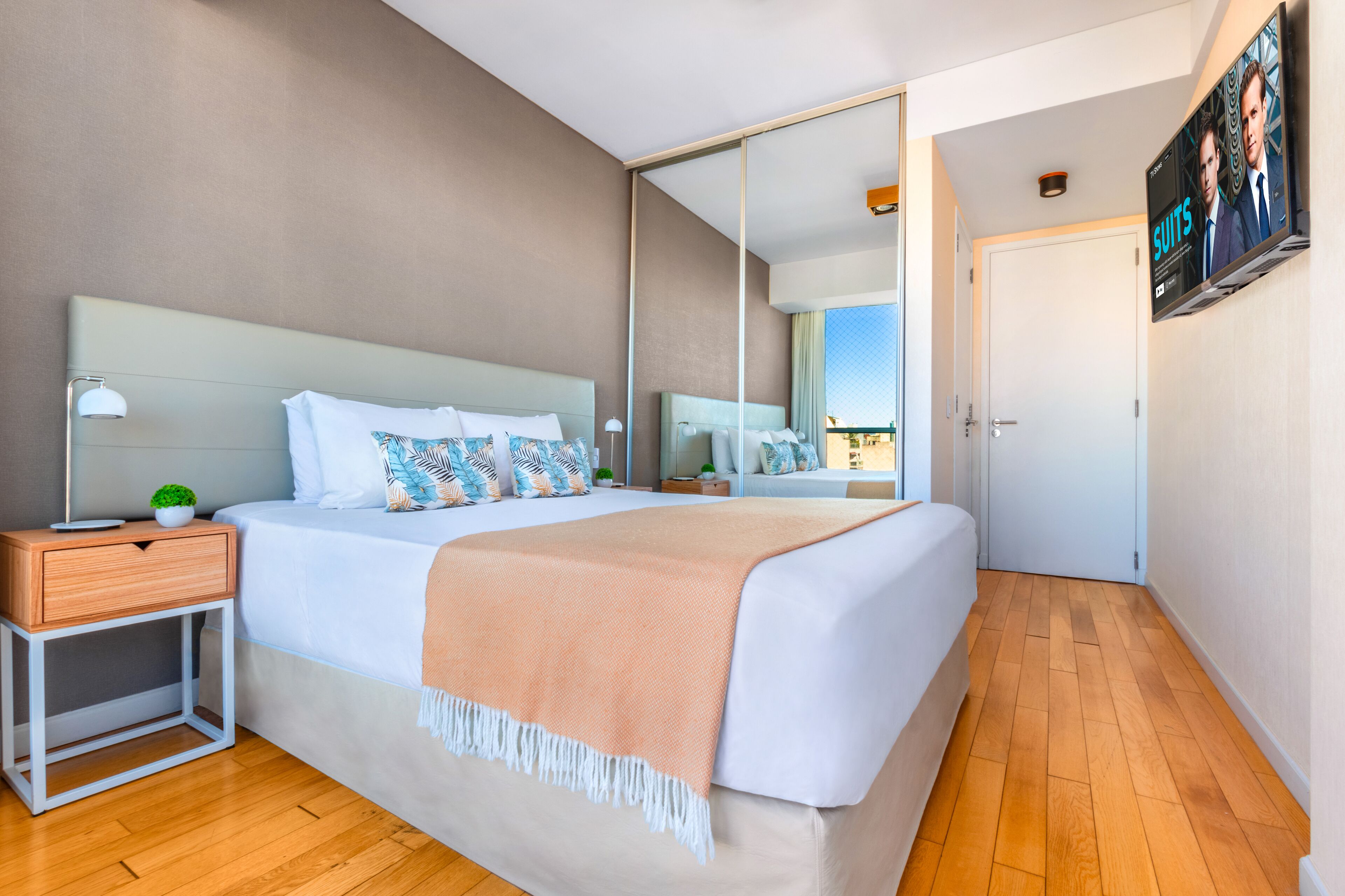Una habitación de hotel moderna con una cama grande, una mesita de noche con lámpara, una planta pequeña y un televisor montado en la pared.