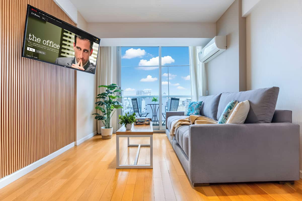 A modern living room with a grey sofa, a wooden floor, a TV on the wall, and a large window with a city view.