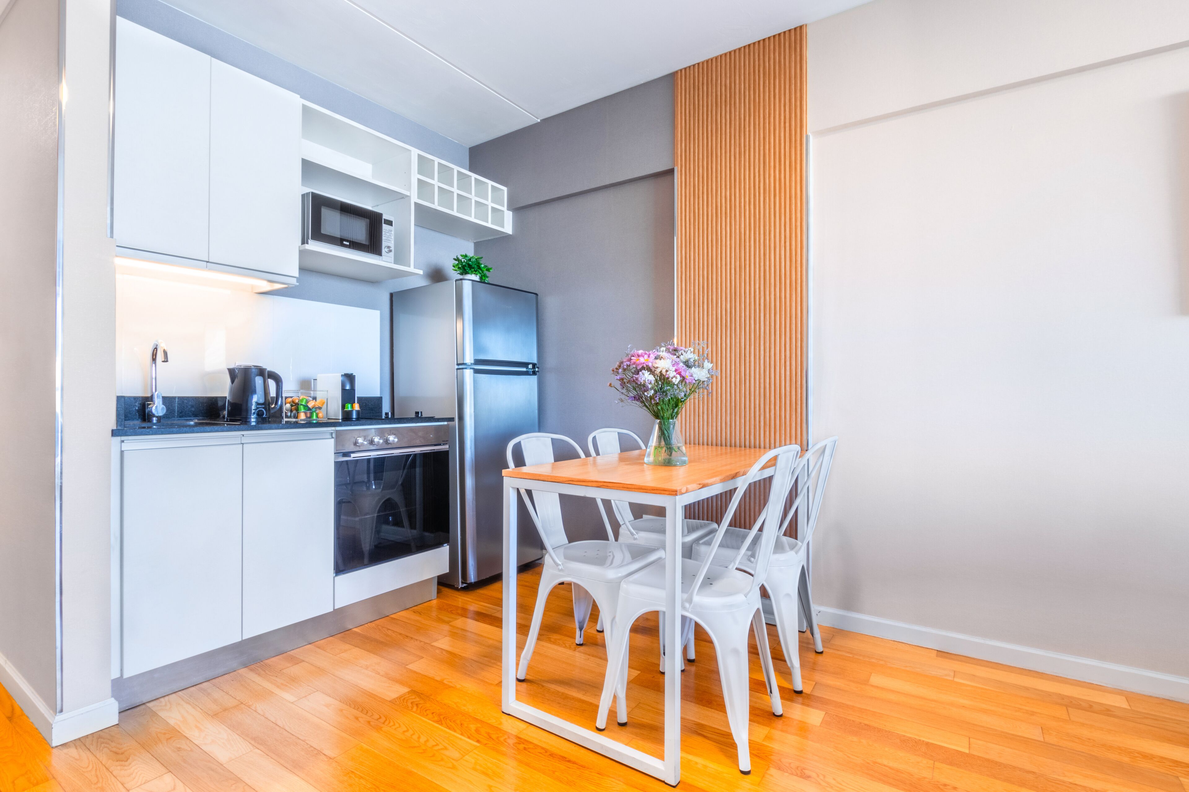 Een compacte keuken met witte kastjes, een roestvrijstalen koelkast, een magnetron en een eethoek met een houten tafel en witte stoelen.