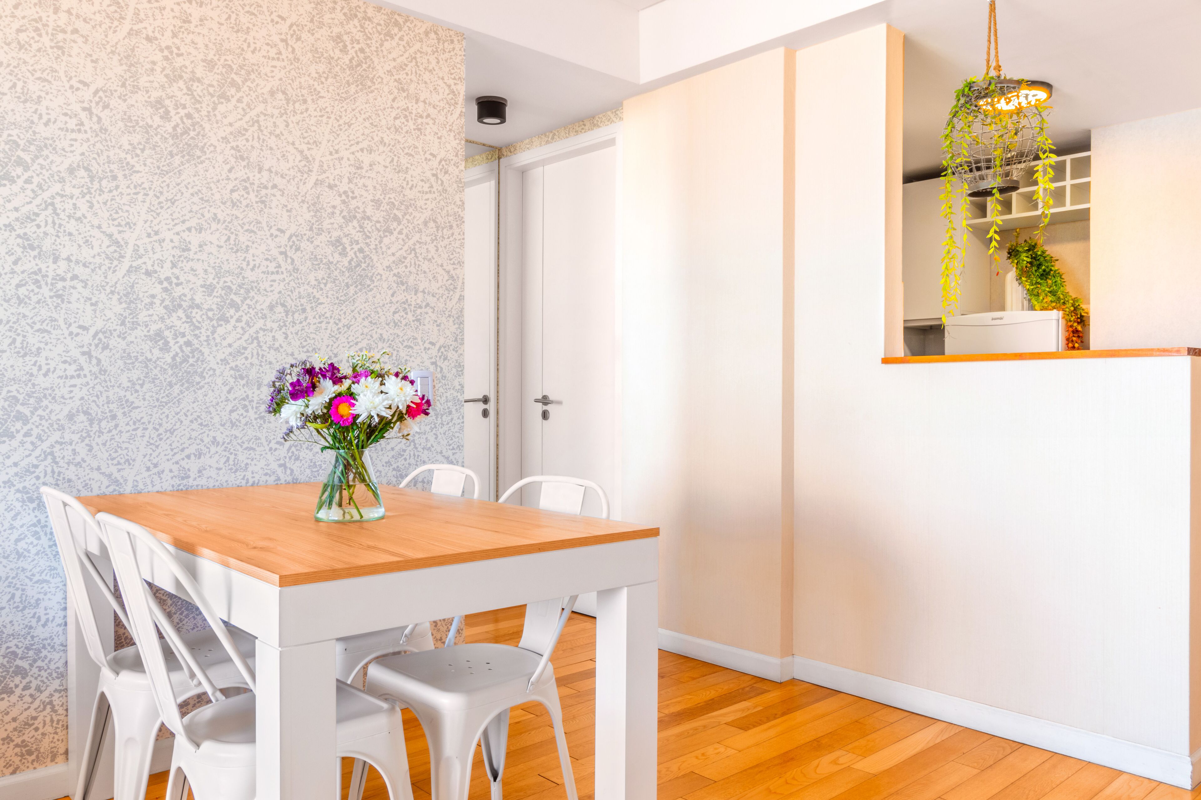 Un comedor con una mesa de madera y sillas blancas, un florero con flores y una planta colgante.