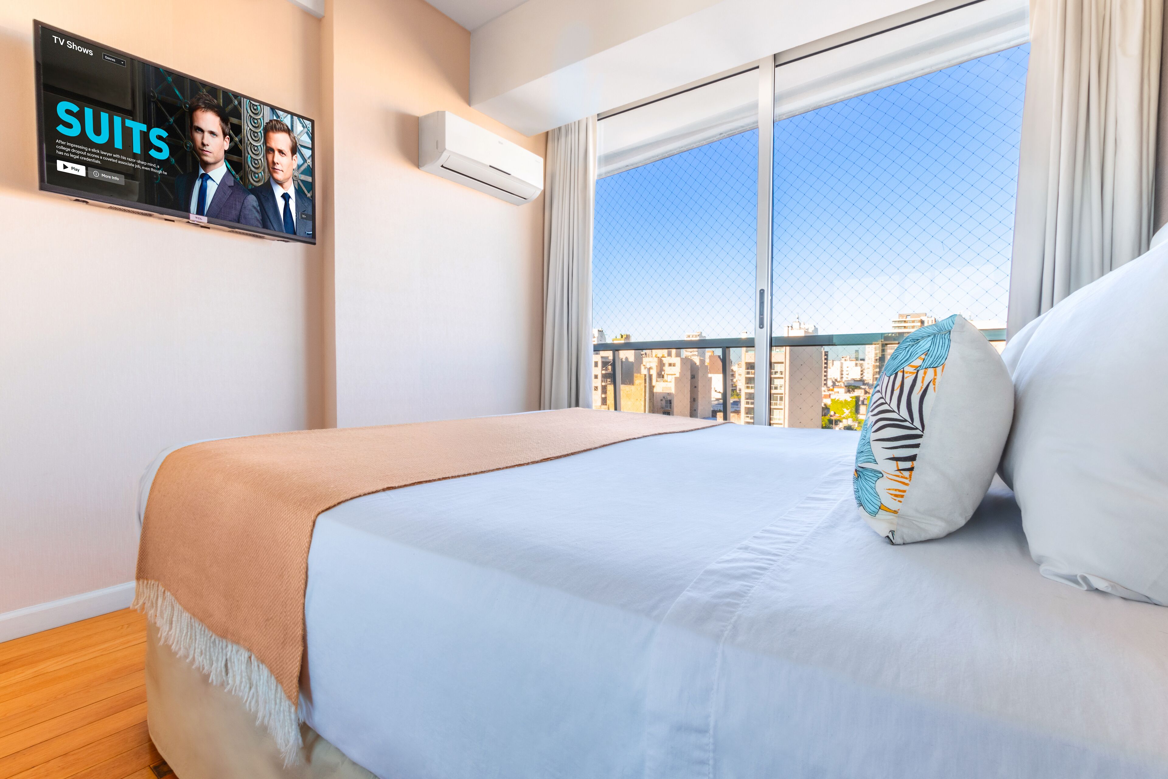 Hotelkamer met een groot bed, een aan de muur bevestigde televisie waarop een programma draait, en een stadszicht door het raam.