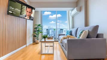 A modern living room with a grey sofa, a wooden floor, a TV on the wall, and a large window with a city view.