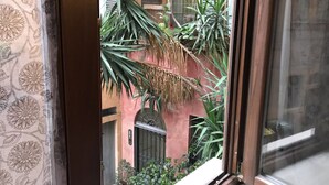 A view through a window with a patterned curtain, featuring a courtyard with potted plants and a building with a red facade and a metal gate.