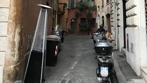 A narrow street with parked scooters and a metal ladder on the left. Buildings with balconies and windows line the street.