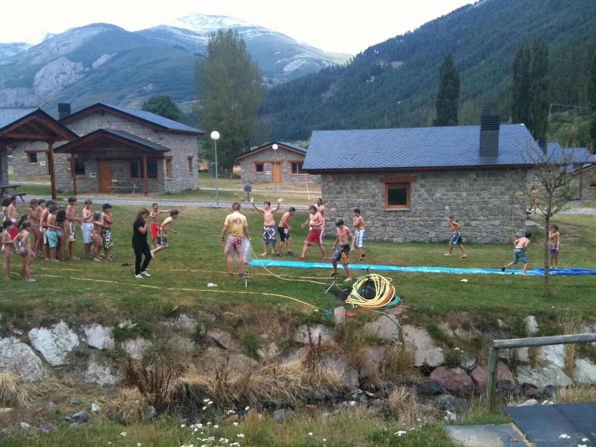 Um grupo de pessoas brincando perto de uma piscina em uma região montanhosa, com edifícios de pedra ao fundo.