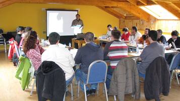 Un salón de conferencias con vigas de madera, una pantalla de proyector y asistentes sentados en mesas con botellas de agua.