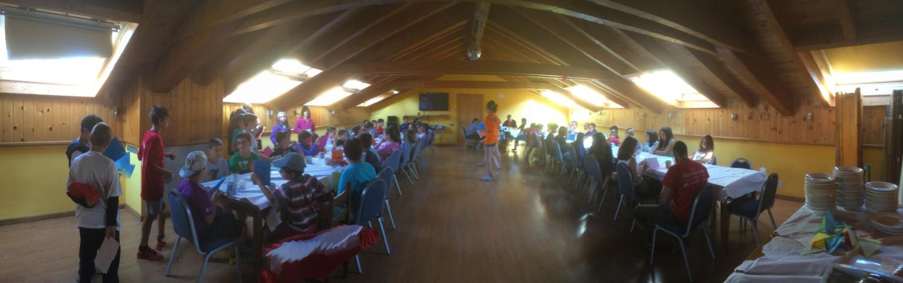 A large hall with high wooden ceilings and natural light from skylights. Tables are set up for a group event.
