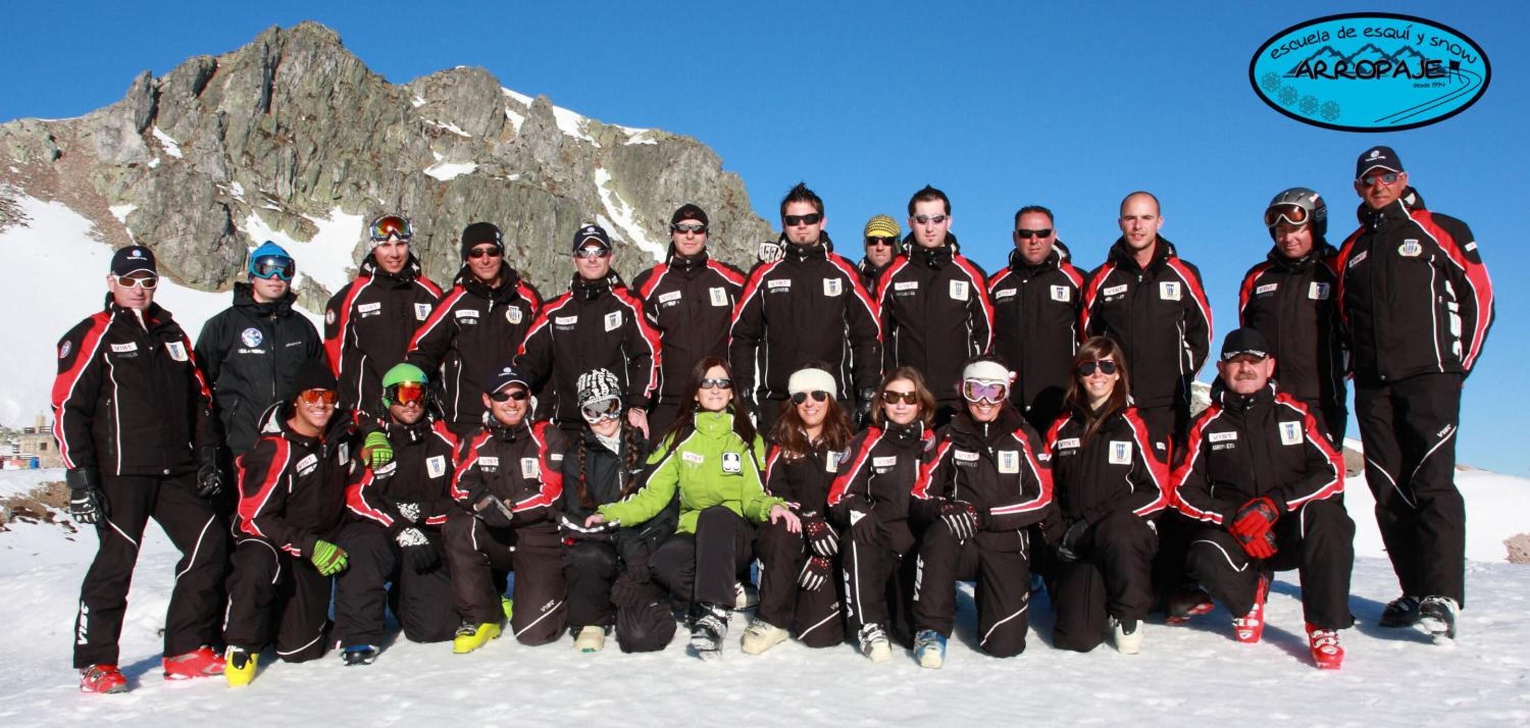 A group of people in winter sports attire posing on a snowy mountain with a rocky backdrop.