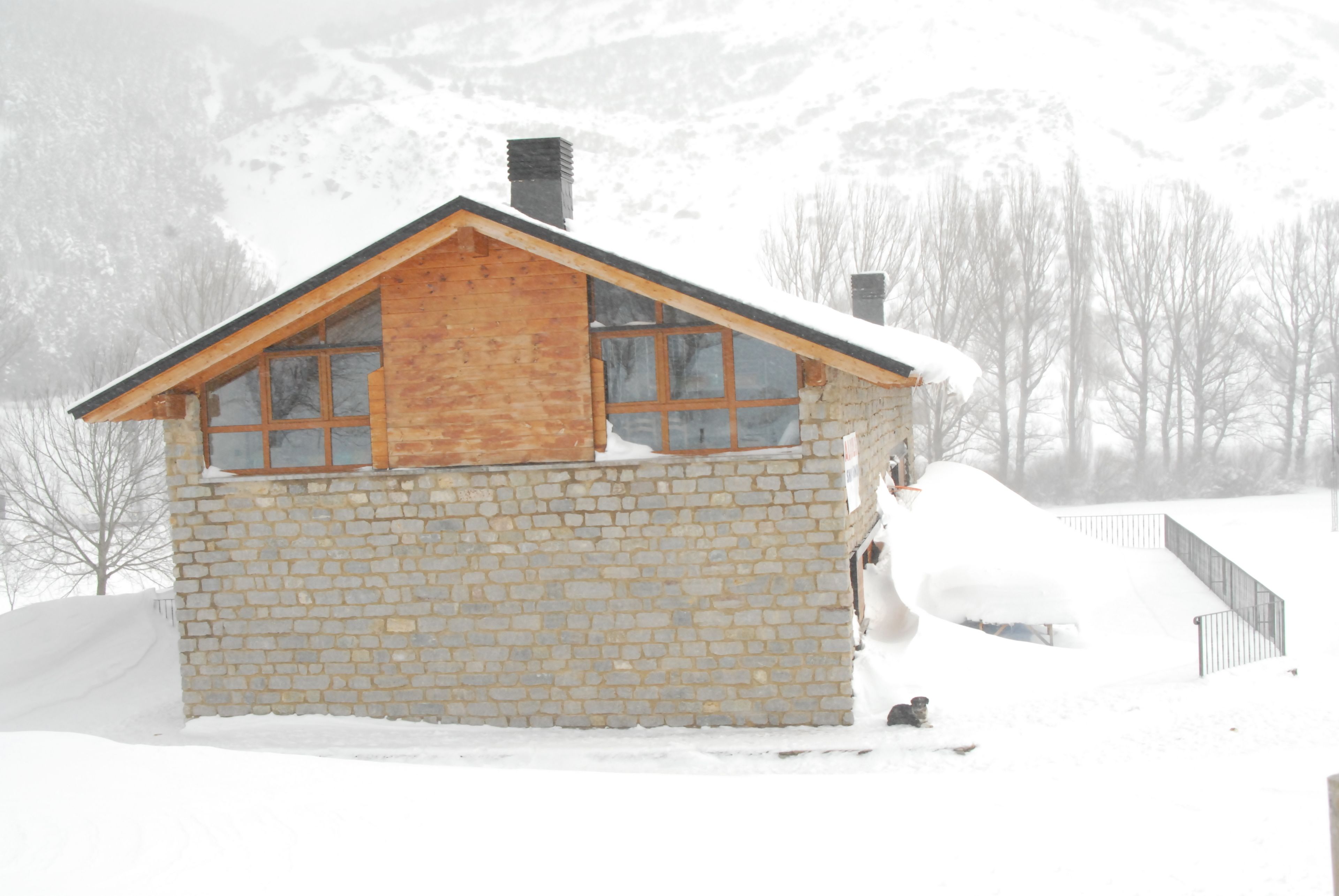 A stone house with a wooden roof and large windows, surrounded by snow-covered mountains.