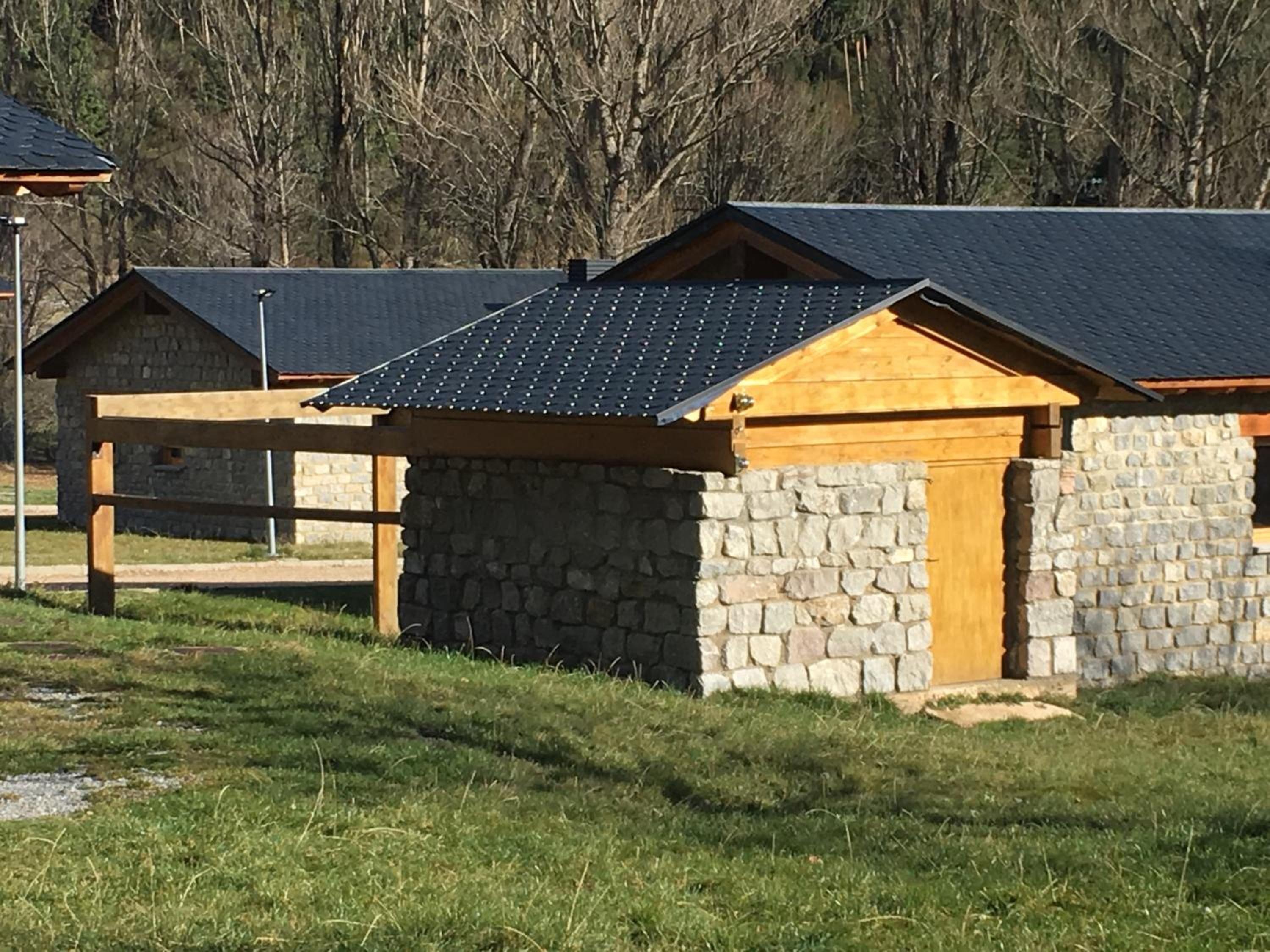 A row of cabins with stone walls and wooden roofs, set in a grassy area with trees in the background.