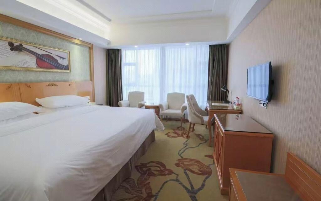 A hotel room with a large bed, a desk, two chairs, a TV, and a window with curtains.
