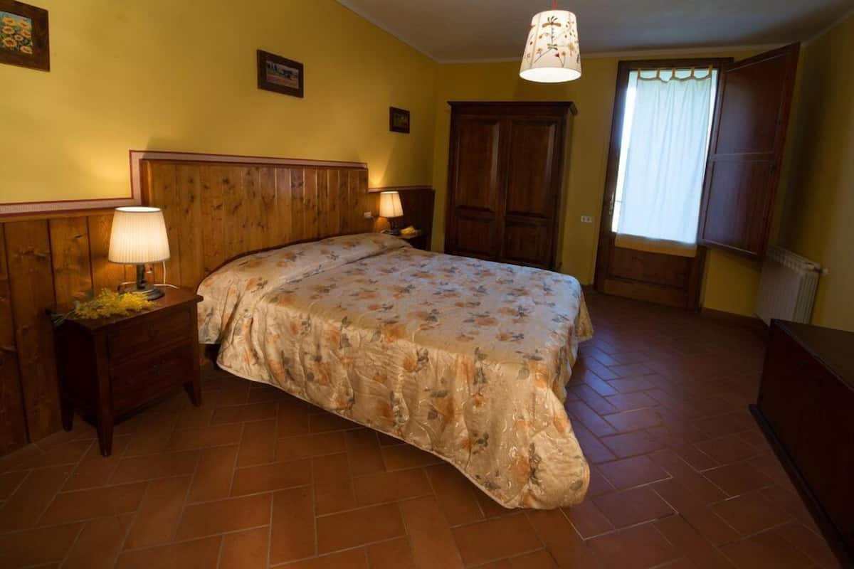 A bedroom with a wooden headboard, a floral bedspread, two bedside lamps, a wooden nightstand, and a large wardrobe.