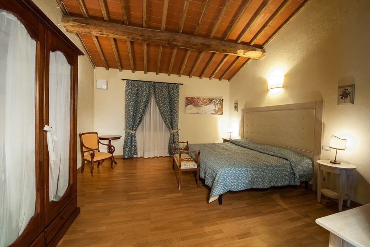 A bedroom with a wooden ceiling, a bed with a patterned bedspread, a chair, a small table, and a nightstand with a lamp.