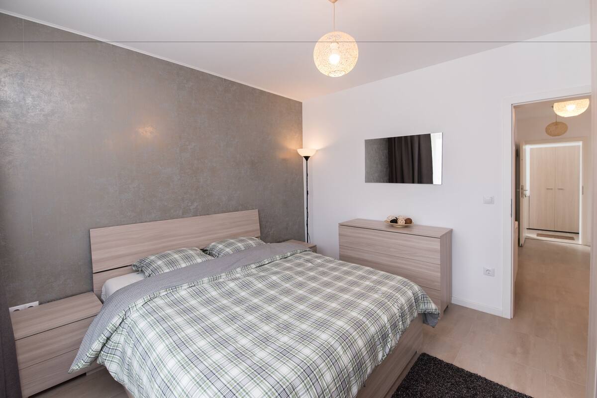 A modern bedroom with a large bed, a bedside table, a nightstand, a mirror, and a door to another room.