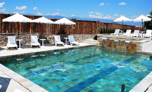A pool with clear blue water, surrounded by white lounge chairs and umbrellas, with a stone wall and mountains in the background. - Hotel Complex Zara Bansko (Bansko)