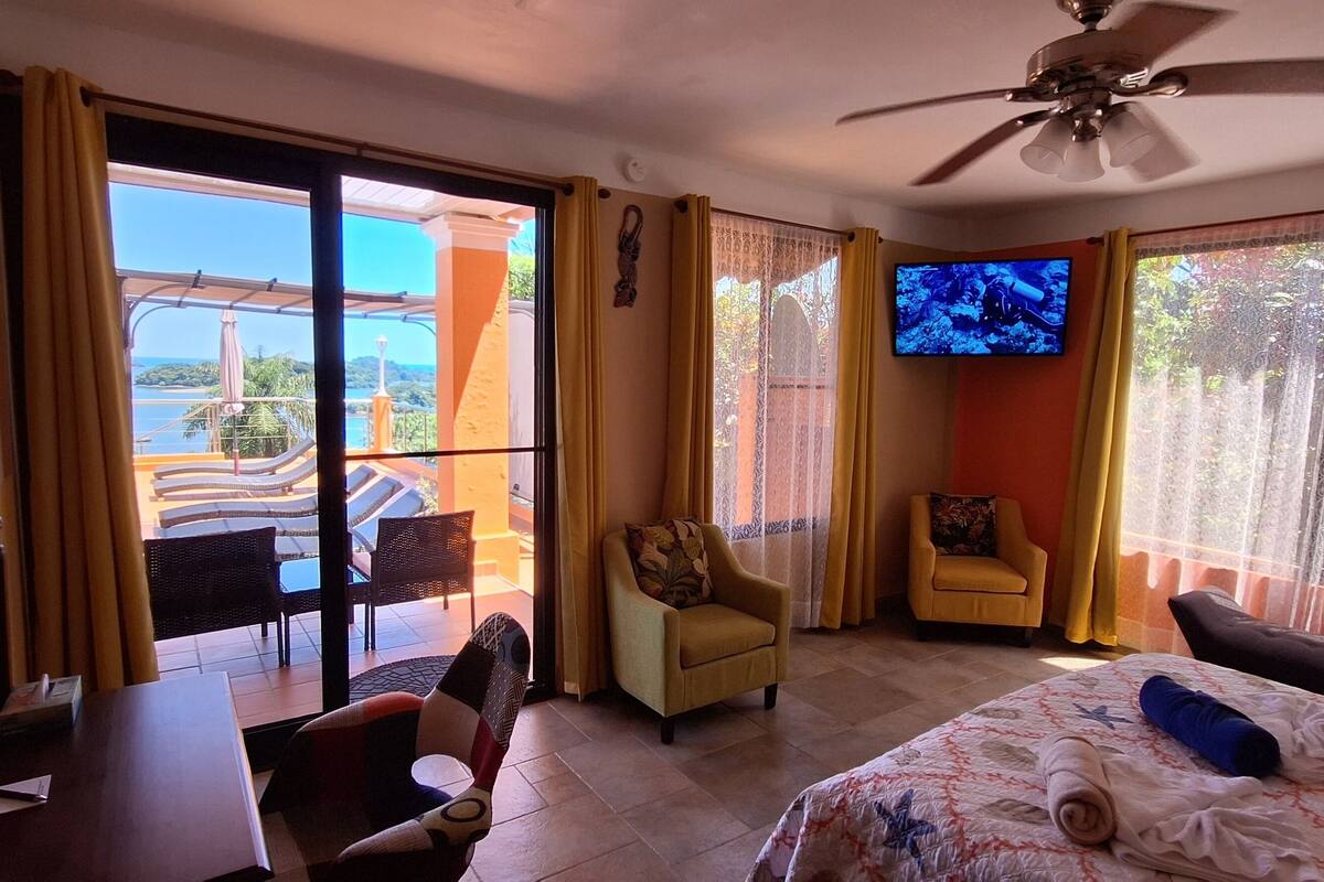 Een hotelkamer met een bed, bureau, stoel, televisie en een balkon met uitzicht op zee.