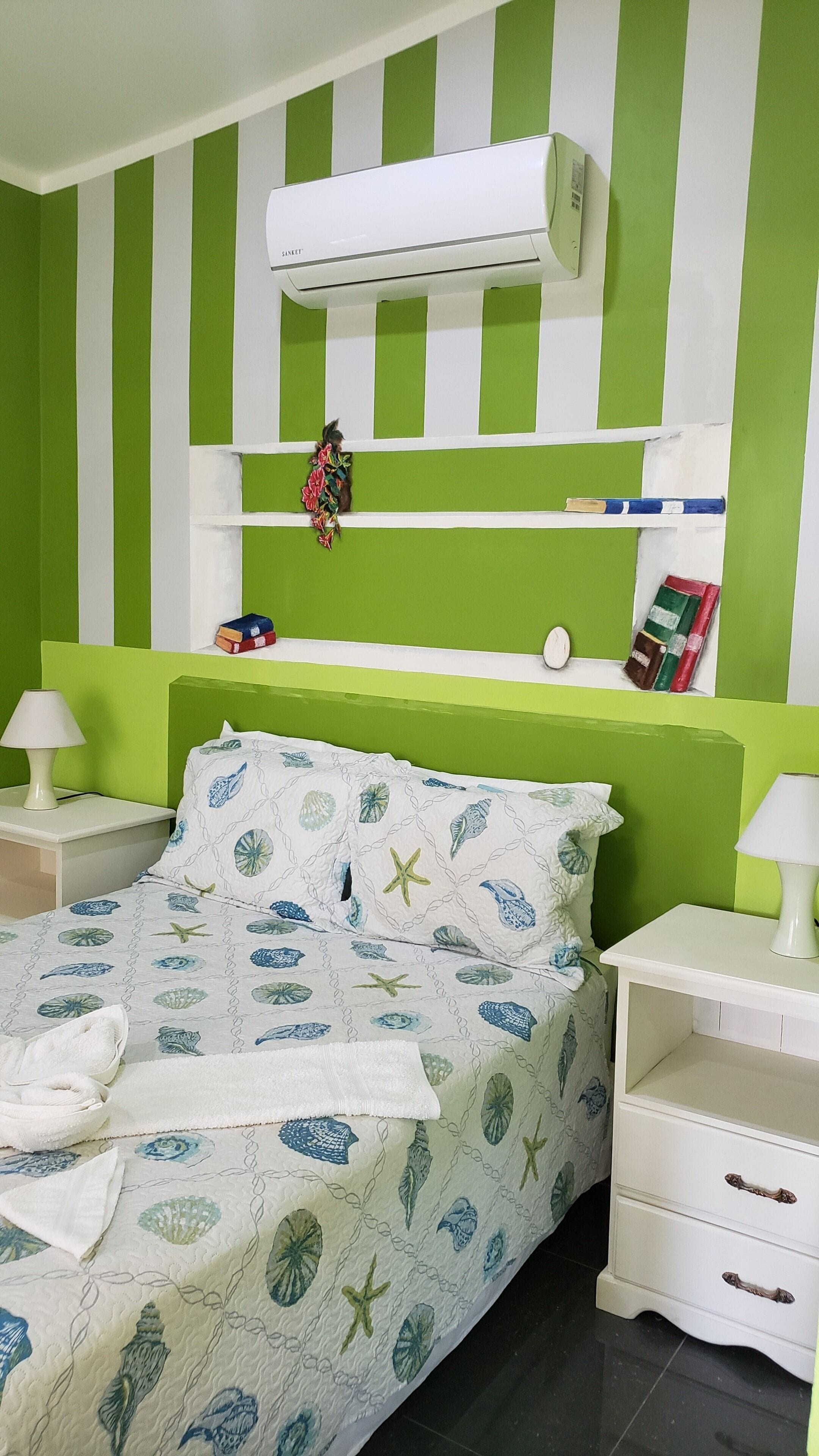 A bedroom with a striped green and white wall, a bed with a leaf-patterned duvet, a white bedside table with a lamp, and a white air conditioning unit above the bed.