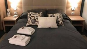 Un lit bien fait, recouvert d’une couvre-lit sombre, avec deux oreillers blancs et un coussin à motifs floraux. Deux tables de chevet avec des lampes sont placées de chaque côté du lit.