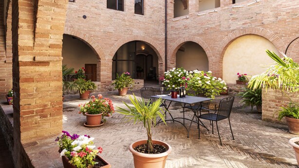 Un patio con muros de ladrillo, arcos, plantas en macetas, una mesa con sillas y una botella de vino.