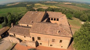 Un edificio grande e histórico con varios niveles y techo de tejas, rodeado de campos verdes y árboles frondosos.