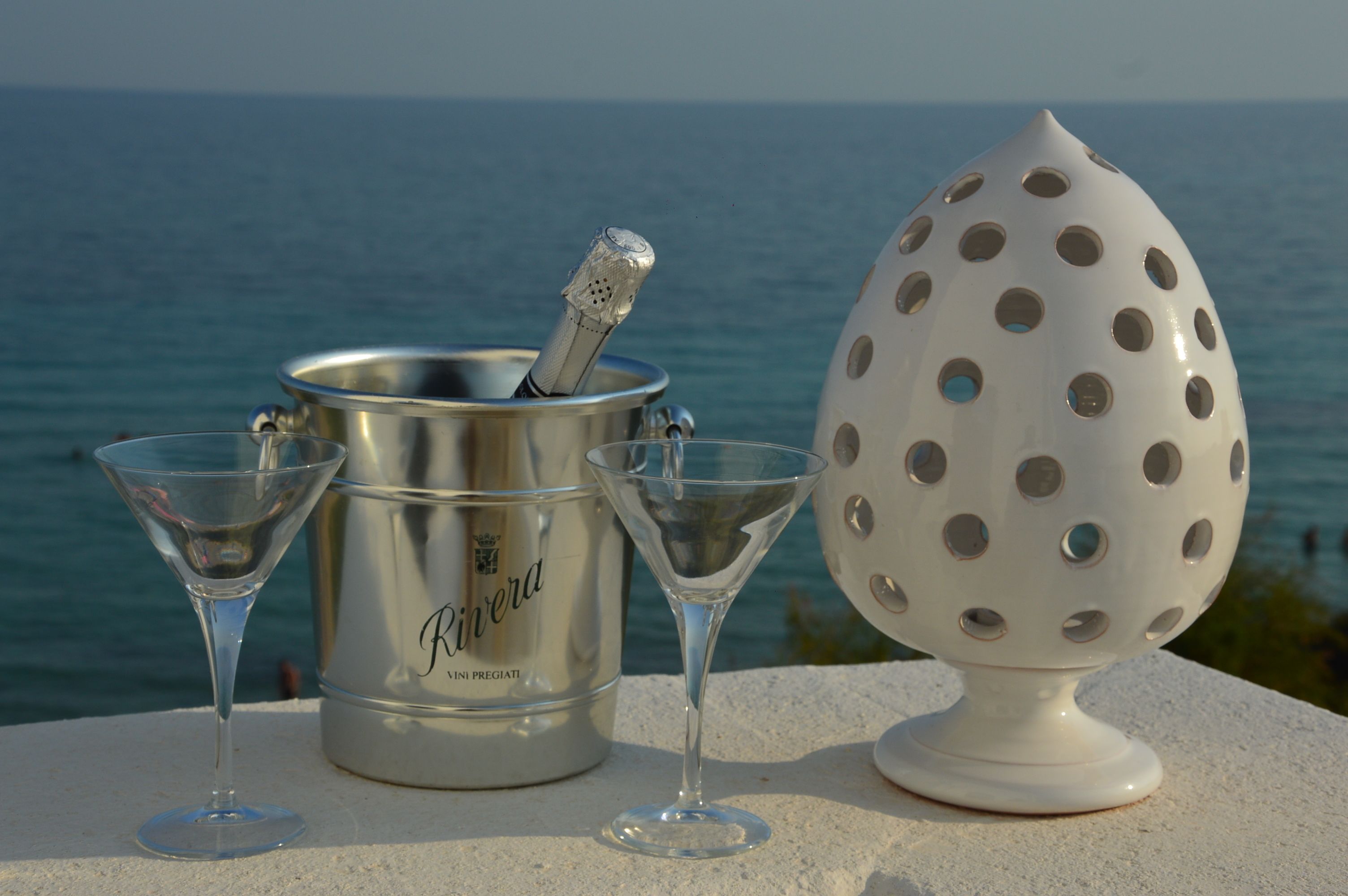 A metallic bucket with a bottle opener and two martini glasses on a ledge with a view of the sea.