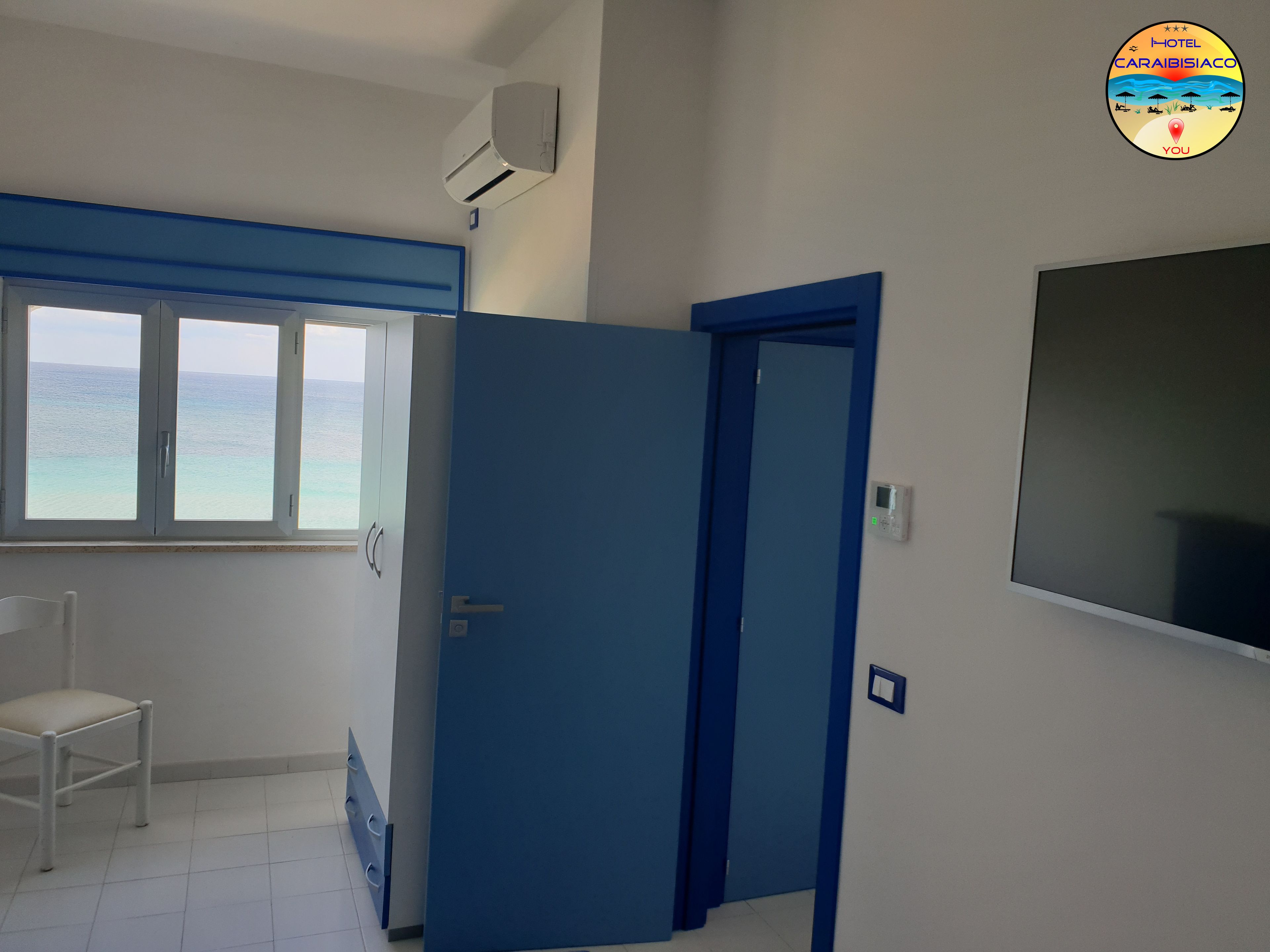 A room with a large window overlooking the sea, a blue door, a white chair, and a wall-mounted air conditioner.